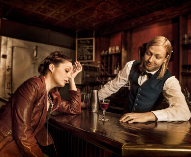 Woman in red jacket leans on bar counter, bartender listens, cocktail between them.