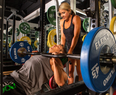 A woman assists a man performing a bench press in a gym, surrounded by colorful weight plates and mirrored walls. Woman spotting man bench pressing in gym with colorful plates and mirrors