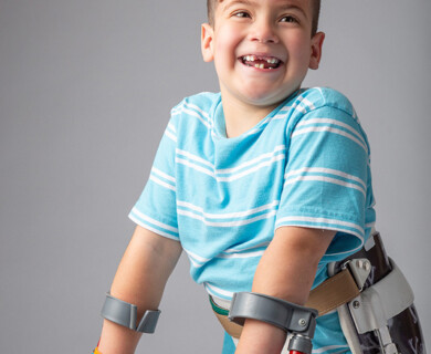 A young boy with red hair smiles broadly while using forearm crutches. He is wearing a blue striped shirt and has colorful wristbands. The background is neutral gray. Young boy with red hair smiling, using forearm crutches, wearing a blue striped shirt.