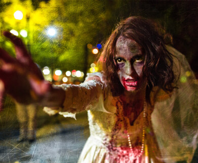 Zombie-costumed person with face makeup reaching forward in dim nighttime setting.