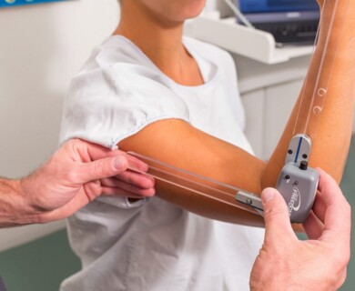 A person is having their arm measured using a medical device with transparent extensions for assessing joint movement. The setting appears to be a clinical or therapy environment, with a computer workstation visible in the background. Person's arm measured using joint movement device in clinical setting.