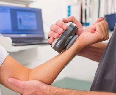 A person is using a medical device to measure or test the strength of another person's arm. A laptop is visible in the background, likely used for data recording or analysis. Person using a dynamometer to test arm strength with data recording on a laptop in the background.