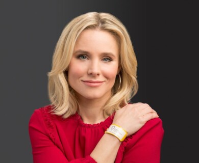 A person with shoulder-length blond hair is wearing a red blouse and a yellow wristband. The individual is smiling softly against a solid dark background. Person with blond hair, red blouse, yellow wristband, smiling against dark background.