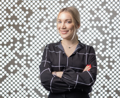 A woman with blonde hair is smiling and standing with arms crossed in front of a backdrop with a pattern of circular lights. She is wearing a black shirt with a white grid pattern. Woman with blonde hair smiling, arms crossed, black shirt, circular light pattern backdrop.