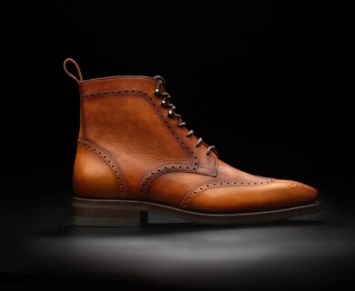 A single brown leather brogue boot is displayed against a black background, featuring detailed decorative perforations and a lace-up style. The boot has a low heel and a pull tab at the back. Brown leather brogue boot with decorative perforations on black background.