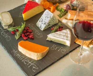 A slate cheese board features a selection of cheeses labeled as Manchego, Montenebro, Rosenborg, Brie, and Mimolette. The board is garnished with dried cranberries and rosemary. Two glasses of red wine and a variety of crackers and bread are placed beside the board. Cheese board with Manchego, Montenebro, Rosenborg, Brie, Mimolette; wine, crackers, garnishes.