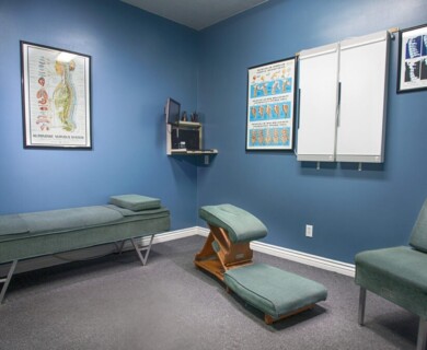 A chiropractic treatment room features a blue color scheme with posters of the nervous system on the walls. The room includes an adjustment table, a specialized chiropractic chair, a regular chair, and a small desk with a computer mounted on the wall. Chiropractic room with blue walls, posters, adjustment table, chair, desk, and wall-mounted computer.