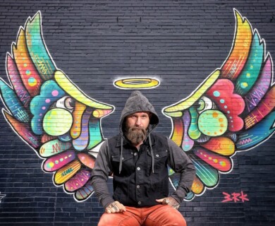 A person in a hooded jacket poses in front of a vibrant mural on a brick wall, featuring stylized, colorful wings with a halo above. Person in hooded jacket in front of vibrant mural with stylized colorful wings and halo.