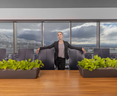 A person stands confidently in a modern conference room with a large wooden table and black chairs. Two planters with green foliage are on the table. Large windows provide a view of a mountainous landscape under a cloudy sky. Person in conference room with wooden table, black chairs, planters, and mountain view.