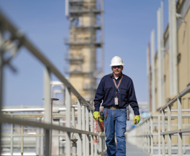 A construction worker is walking on a metal walkway, wearing a white hard hat, blue uniform, and yellow gloves. The background features an industrial setting with metal structures and equipment. Construction worker on metal walkway in hard hat, blue uniform, yellow gloves, industrial background.