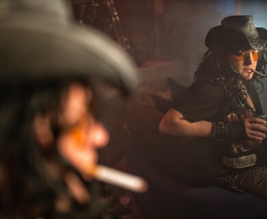 A person wearing a cowboy hat and yellow-tinted glasses is sitting in a dimly lit room. They have a cigarette in their mouth and are holding a drink, dressed in eclectic clothing including zebra-patterned pants. The scene has a smoky and moody atmosphere. Person in cowboy hat, yellow glasses, smoking, holding drink in smoky, moody room.