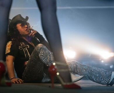 A person wearing a cowboy hat, sunglasses, and zebra-patterned pants is sitting on the ground, smoking. Red high heels and legs in stockings are visible in the foreground, and car headlights illuminate the background. Person in cowboy hat and zebra pants sitting, smoking, with car headlights in the background.