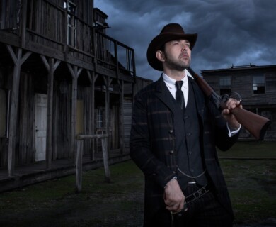 A man wearing a suit and tie, with a cowboy hat, holds a rifle while standing in front of an old, wooden building. The sky is overcast, adding a dramatic atmosphere to the scene. Man in cowboy hat with rifle, standing by old wooden building under overcast sky.