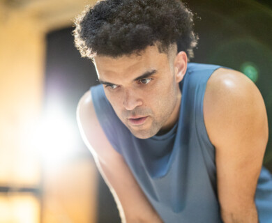 A person with short curly hair is leaning forward, wearing a sleeveless top. The lighting is bright, with lens flare visible in the background. Person with short curly hair in sleeveless top, bright light with lens flare behind them.
