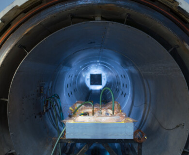 The image depicts the interior of a large, cylindrical chamber, possibly a wind tunnel or industrial machinery. Inside, there is equipment connected by several tubes and wires, and the entire setup is lit with a blue tone. Interior of a cylindrical chamber with machinery, wires, tubes, and blue lighting.