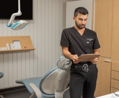 A dental professional in black scrubs is standing in an examination room, reading a clipboard. A dental chair and overhead light are visible, with a wooden shelf and some cabinets in the background. Dental professional in scrubs reads clipboard in exam room with chair, light, and cabinets.