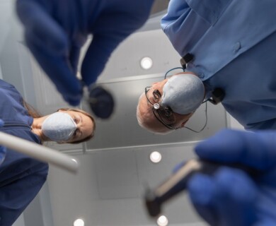 Two dental professionals wearing masks and gloves are seen from the perspective of a patient. They are holding dental instruments, likely preparing for a procedure. Two masked dental professionals with instruments, viewed from patient perspective in a clinic.