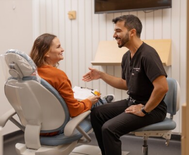 A dentist in black scrubs is smiling and engaging in conversation with a female patient seated in a dental chair. The setting appears to be a dental clinic, with a clipboard visible in the patient's hands. Dentist in black scrubs converses with a smiling female patient in a dental clinic.