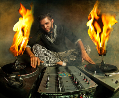A person is sitting cross-legged behind a DJ setup with two turntables and a mixer. Both turntables have records on them with flames emerging from the center, creating a dramatic visual effect. The individual is wearing patterned pants and a dark top, and appears to be engaged in DJing. DJ performing with flaming records on turntables, cross-legged behind mixer setup.