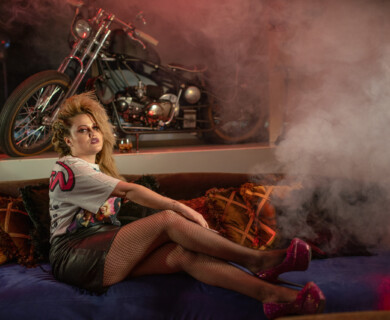 A person with dramatic makeup and voluminous hair is seated on a couch with colorful cushions. They are wearing fishnet stockings, a leather skirt, and a graphic t-shirt. In the background, a motorcycle is visible, and the area is filled with smoke, creating a dynamic atmosphere. Person with dramatic makeup on couch, behind them a motorcycle and smoke-filled atmosphere.