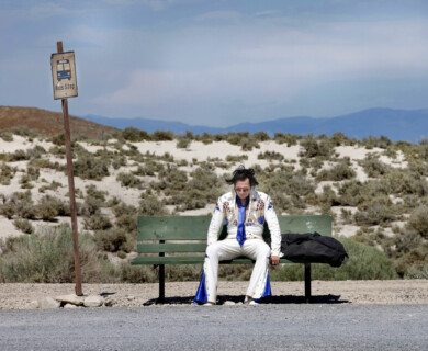 A person dressed in an Elvis Presley-inspired costume is sitting on a bench at a bus stop in a desert landscape. The sign beside them indicates a bus stop, and the background features arid terrain and distant mountains. Person in Elvis costume sitting at desert bus stop with arid terrain and mountains in background.