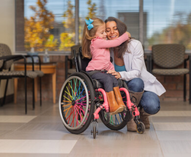 A young girl in a wheelchair with colorful spokes is hugging a woman who is kneeling beside her. They are in a room with windows and chairs in the background. The girl is wearing a pink shirt and brown boots, while the woman is in a white jacket. Young girl in wheelchair with colorful spokes hugging woman in a room with windows and chairs.