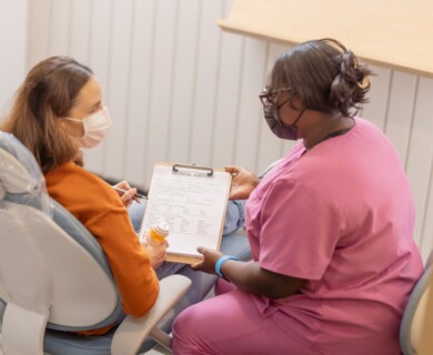 A healthcare professional in pink scrubs is discussing a medical history form with a patient seated in a chair. Both individuals are wearing face masks. The patient holds a bottle of medication while engaging with the form. Healthcare worker in pink scrubs discusses medical form with masked patient holding medication.