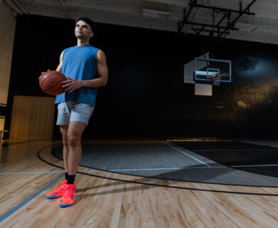 A person is standing on an indoor basketball court holding a basketball. They are wearing a sleeveless blue shirt, gray shorts, and bright orange athletic shoes. The court is well-lit, with a basketball hoop visible in the background. Person on indoor basketball court holding ball in blue shirt, gray shorts, orange shoes.