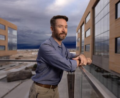 A man with a beard is standing on a balcony, leaning on the railing, with a modern building and overcast sky in the background. Man with beard on balcony, leaning on railing with modern building and overcast sky behind.
