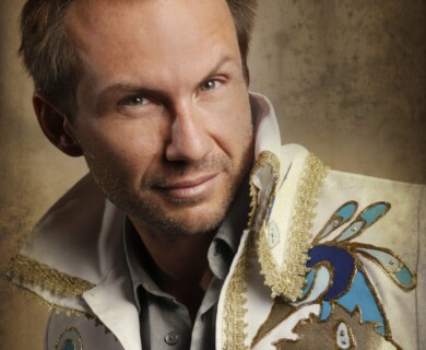The image depicts a man with short hair wearing a decorative jacket featuring intricate patterns in blue and gold. The background is a textured, neutral shade. Man in a blue and gold patterned jacket against a textured neutral background.
