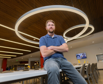 A man in a blue shirt is sitting on a table in a modern interior space with circular ceiling lights and wooden paneling. Several chairs and tables are visible in the background, indicating a possible cafeteria or communal area. Man in blue shirt seated on table in modern room with circular lights, wood paneling, chairs.