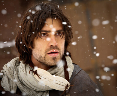 A man with shoulder-length brown hair and a beard, wearing a light-colored scarf and a decorative mask around his neck, is standing amidst falling snowflakes. Man with brown hair and beard in snow, wearing light scarf and mask around neck.