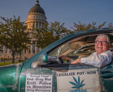 A man is sitting in a vehicle decorated with signs promoting the legalization of cannabis and addressing social issues. The vehicle is parked near a large domed government building. Man in vehicle with signs for cannabis legalization near large domed government building.