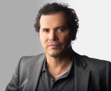 A man with short, curly hair is wearing a dark blazer over a collared shirt and a silver necklace. The background is split between gray and white. Man with curly hair in blazer, collared shirt, silver necklace; gray and white background.