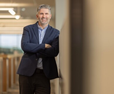 A man with gray hair is leaning against a wall in an office environment. He is wearing a navy blue blazer over a light blue shirt and has a friendly expression. The background shows rows of office cubicles and overhead lighting. Man with gray hair in blue blazer in office, wall-leaning with cubicles in background.