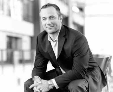 A man in a suit is seated, leaning slightly forward, in a bright, modern interior environment. The image is in black and white, with a blurred background suggesting a professional setting. Man in suit seated indoors, black and white, professional blurred background.