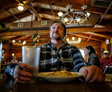 A man in a plaid shirt sits at a wooden table in a rustic restaurant with wooden beams and decorative lighting. He is holding a cup and has a plate of food in front of him. Other patrons are visible in the background. Man in plaid shirt holding a cup at a rustic restaurant with wooden beams and decorative lighting.