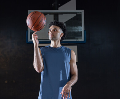 A man is standing indoors, spinning a basketball on his finger. He is wearing a sleeveless shirt, and there is a basketball hoop in the background. The setting appears to be a gym or sports facility. Man indoors spinning basketball on finger, wearing sleeveless shirt, gym hoop in background.