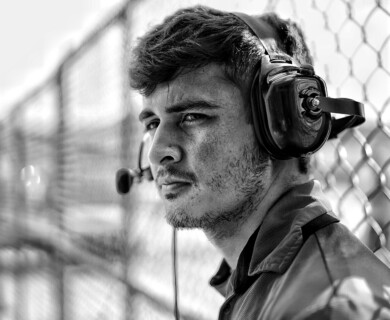 A monochrome image featuring a man wearing a headset with a microphone. He is standing next to a chain-link fence, appearing attentive and focused. Man with headset and mic next to chain-link fence, attentive and focused in monochrome image.