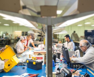 A group of workers in a manufacturing or assembly setting are seated at a workspace. They are focused on their tasks, working with tools and equipment, including cases and boxes. The environment appears organized and industrious, with people wearing safety glasses and lab coats. Workers in a manufacturing setting focused on tasks with tools and safety gear.