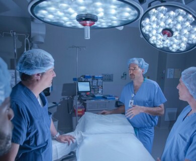 A group of medical professionals wearing surgical scrubs and caps are gathered in an operating room, with one person appearing to explain or discuss something. Bright surgical lights are overhead, and medical equipment is visible in the background. Medical team in scrubs discussing in an operating room with surgical lights overhead.