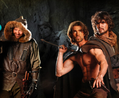 Three men dressed in medieval-style clothing are in a dimly lit cave. One is holding a glowing blue potion, another is shirtless with a dagger drawn, and the third wears a cloak. The setting appears adventurous and mysterious. Three men in medieval attire inside a cave; one holds a blue potion, another wields a dagger.