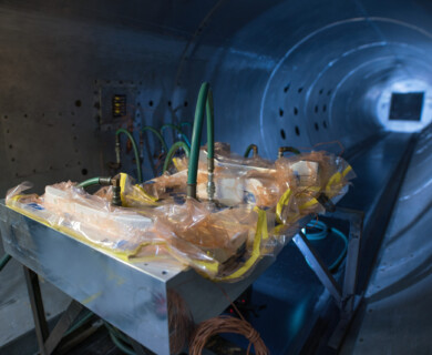 The image shows a metallic structure inside a tunnel, with various green hoses attached to it and surrounded by protective plastic wrapping. The tunnel widens at the far end, suggesting an industrial or testing environment. Metallic structure with green hoses in tunnel, surrounded by plastic wrapping, in industrial setting.