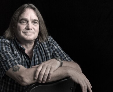 A middle-aged person with shoulder-length hair is seated against a black background, wearing a plaid shirt and displaying a tattoo on their upper arm. Their hands are resting on what appears to be the back of a chair. Middle-aged person in plaid shirt with tattoo on arm, seated against black background.