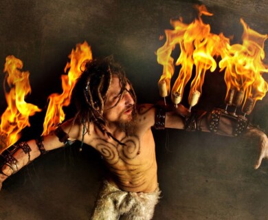 A performer with dreadlocks and tattoos is depicted wielding flaming sticks attached to arm braces. They are dressed in a dramatic costume, including fur-like pants, set against a dark background. Performer with tattoos and dreadlocks holding flaming sticks on arm braces in a dramatic costume.