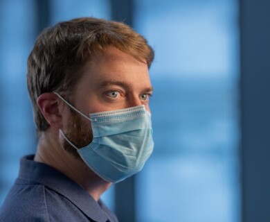 A person with short hair and a beard is wearing a blue surgical mask, looking to the side. The background is blurred and has cool tones, possibly indicating an indoor setting. Person with short hair, beard, wearing blue surgical mask; blurred cool-toned background.
