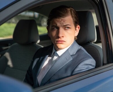A person wearing a blue suit and white shirt is sitting in a car, looking out of the window with a contemplative expression. The car is parked, and there is grass visible outside. Person in blue suit sitting in parked car, looking out window; grass outside.