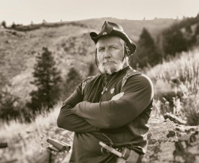 An individual with a beard and wearing a hat stands outdoors in a mountainous area, with arms crossed. The image is in black and white, capturing the natural scenery and rugged terrain in the background. Individual wearing a hat stands with arms crossed outdoors in a mountainous area, black and white.