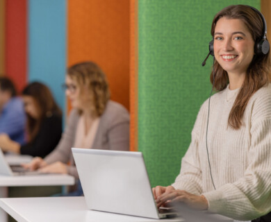 A person wearing a headset is seated at a table working on a laptop, with other individuals focusing on laptops in the background. The setting features colorful partitioned walls. Person with headset at laptop, others working in background, colorful partitioned walls.