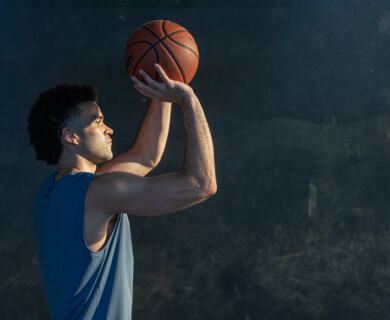 A person is poised to shoot a basketball, holding the ball above their head with both hands. They are wearing a sleeveless shirt and are focused on their action against a plain, dark background. Person holding basketball above head, wearing sleeveless shirt, against dark background.